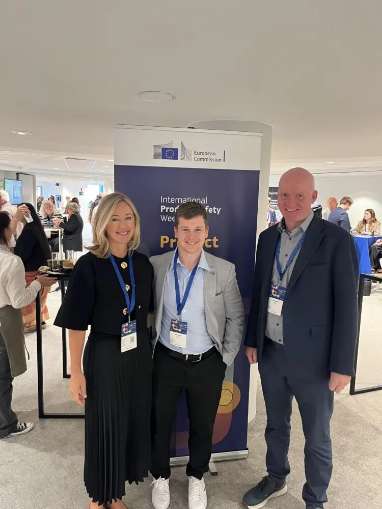 Three smiling people, two men and one woman, stand in front of a European Commission banner at an event. The banner mentions 'International Product Safety Week'.