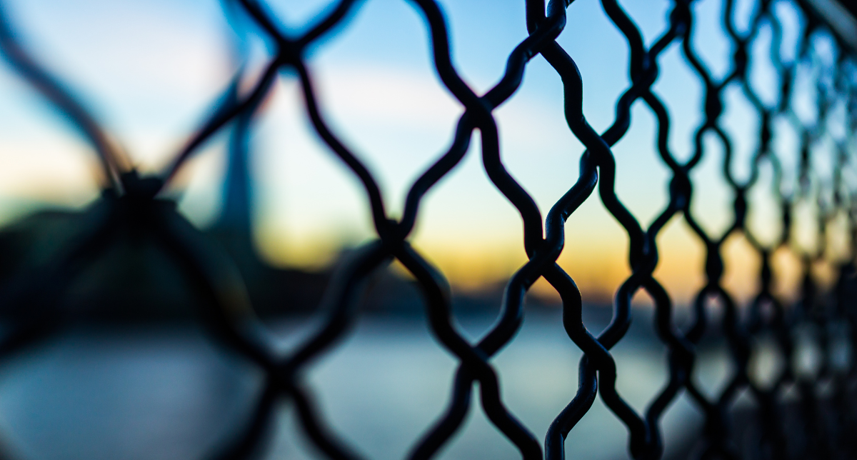 A close-up of a dark chain-link fence in sharp focus, with a blurred background showing a blue sky, orange horizon, and dark water.