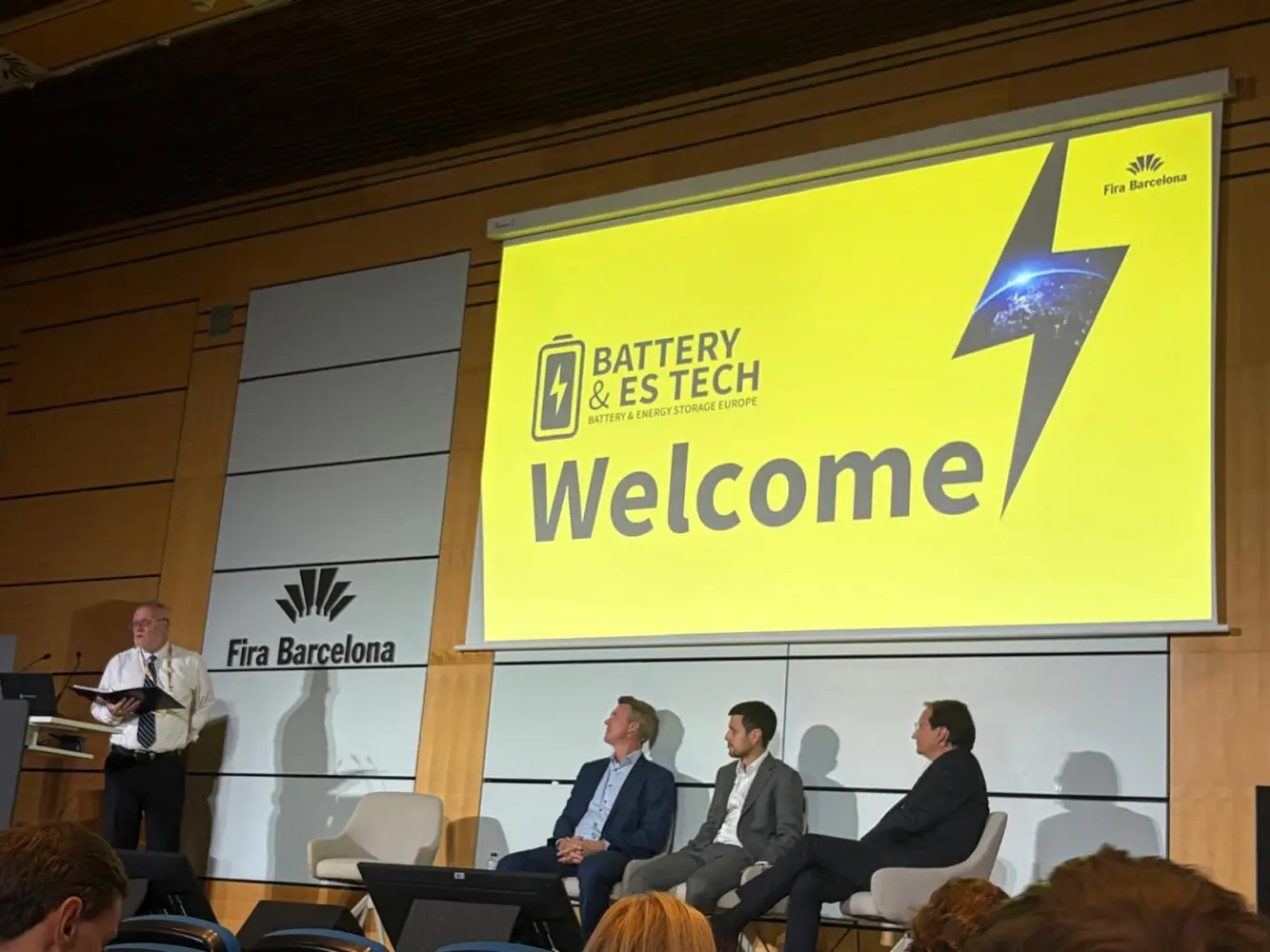A conference stage with a speaker at a podium and three panelists seated, all in front of a large yellow screen displaying 'Welcome' and 'Battery & ES Tech' branding.