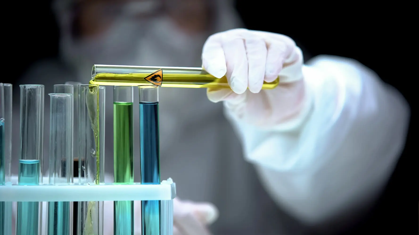 A gloved hand in a protective suit pours yellow liquid from a biohazard-labeled test tube into another, amidst a rack of test tubes with various colored liquids.