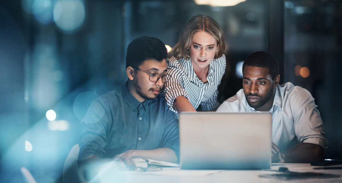 Three diverse colleagues, two men and one woman, intently looking at a laptop screen in a dark office, collaborating late at night.