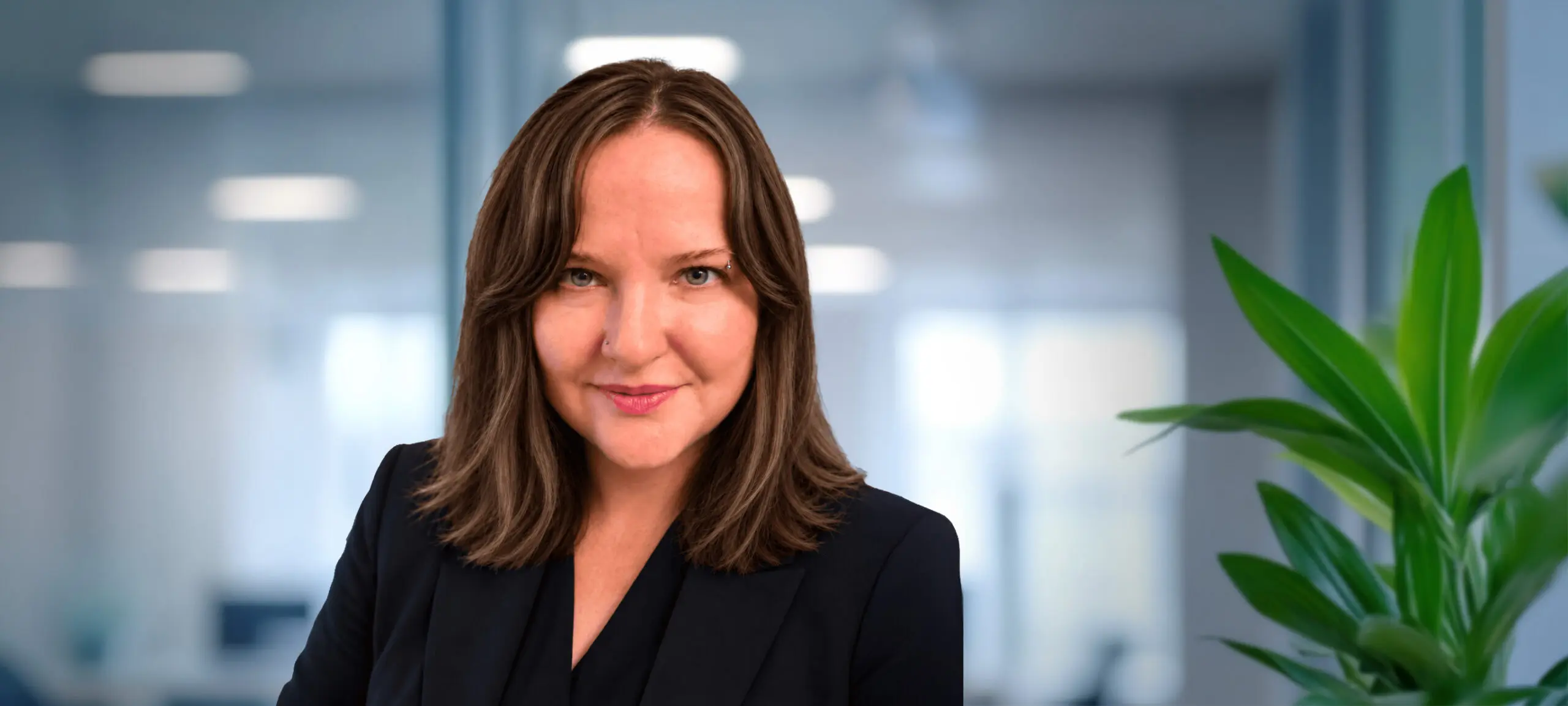 Headshot of a woman with brown hair, blue eyes, and a slight smile, wearing a dark blazer. She has a nose stud and an eyebrow piercing. The background is a blurred office.