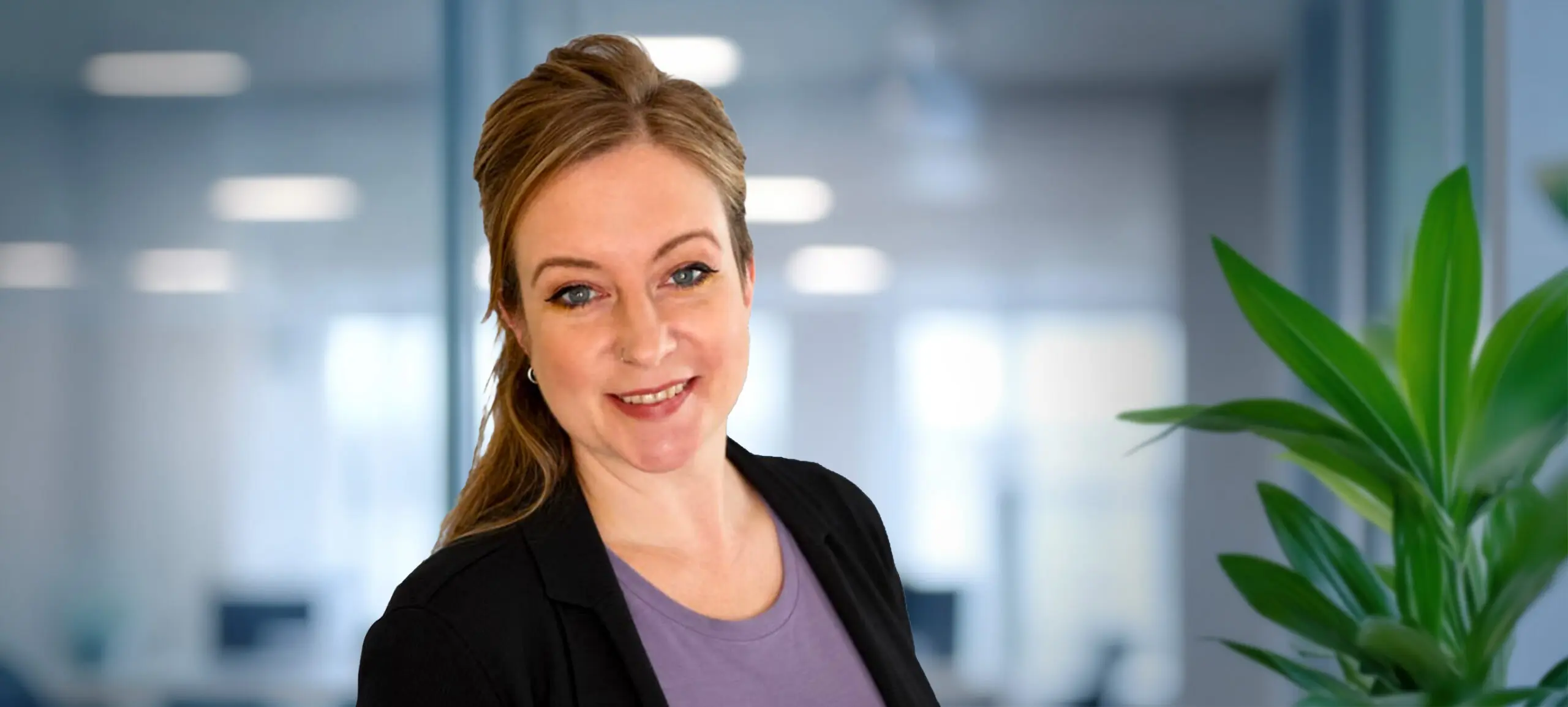 A smiling woman with light brown hair and a black blazer stands in a blurred office setting with a green plant on the right.