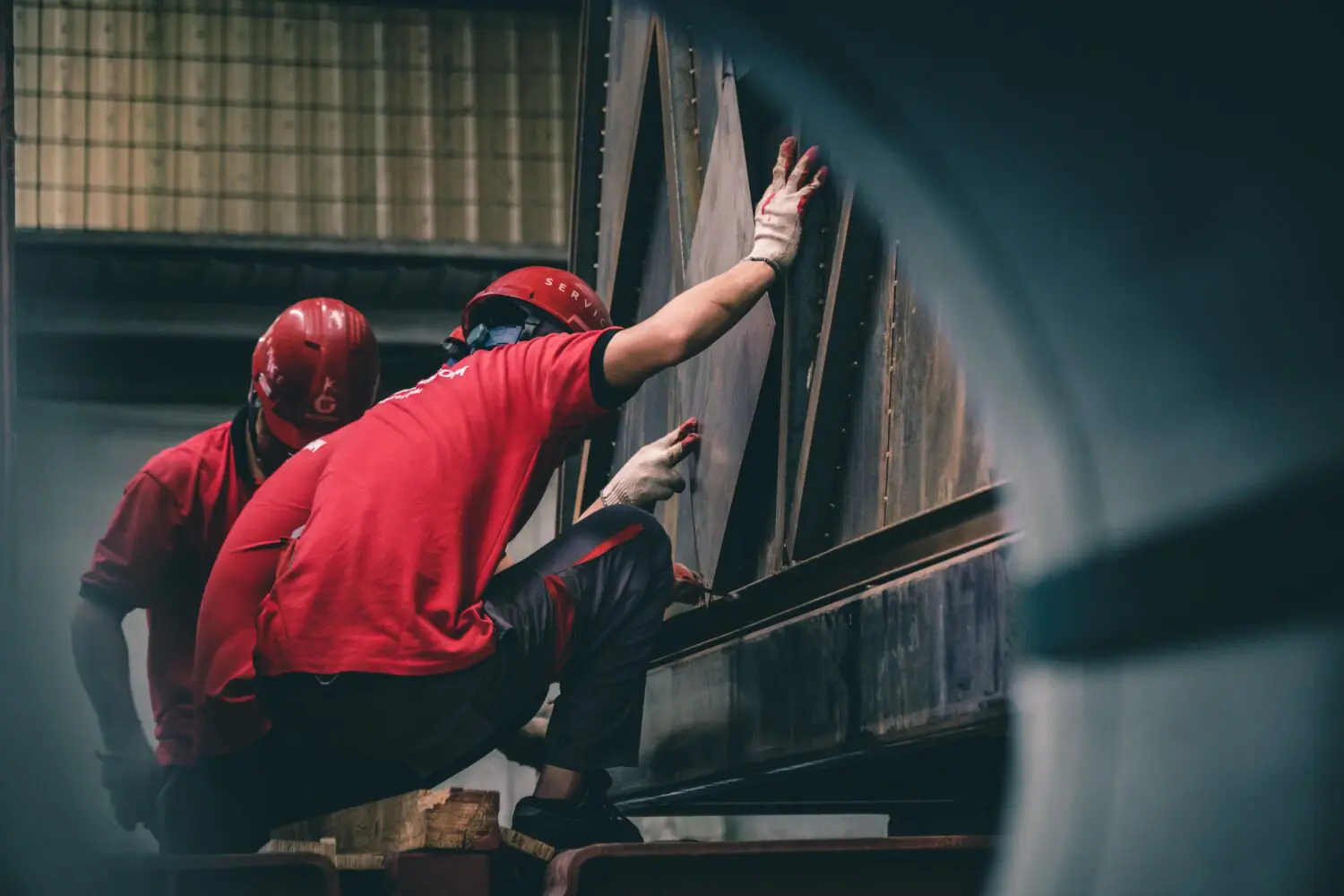 Two workers in red shirts and red hard hats working on a large metal structure in an industrial setting. One worker is touching the metal with a gloved hand.