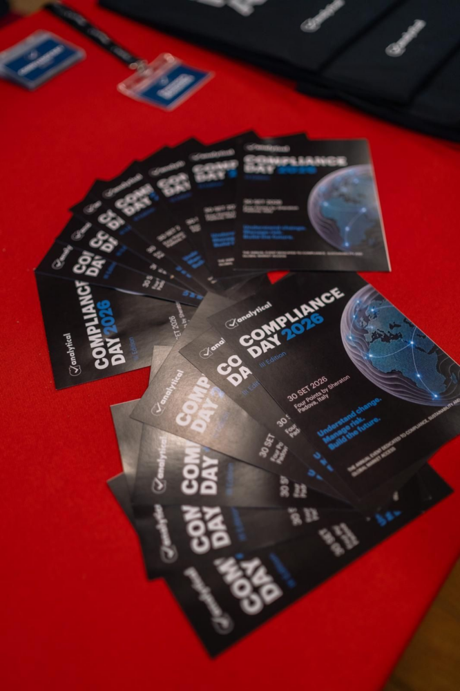A fan of black and blue event flyers for 'Compliance Day 2026' laid on a red table, with out-of-focus badges and merchandise in the background.