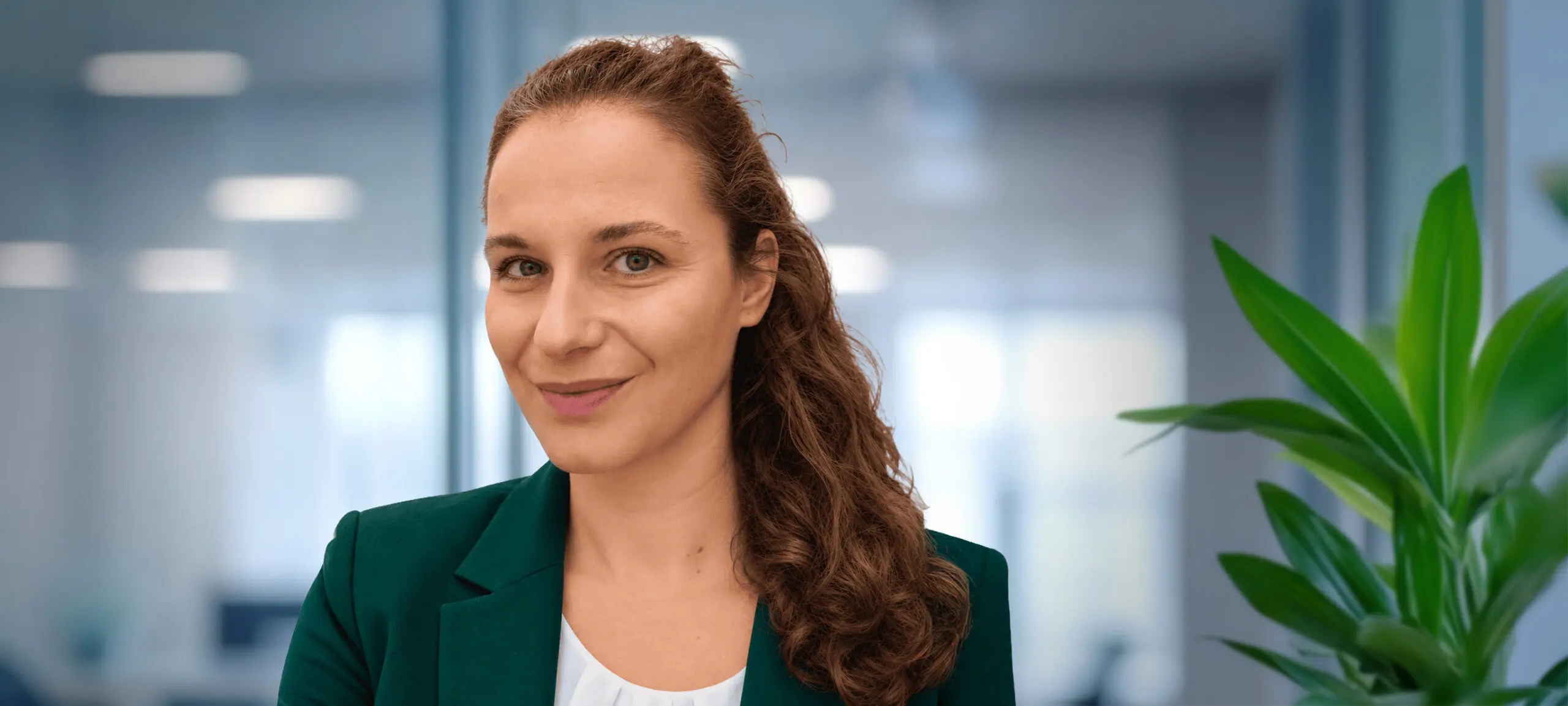 A woman with light brown, wavy hair and a green blazer smiles at the camera in an office setting.