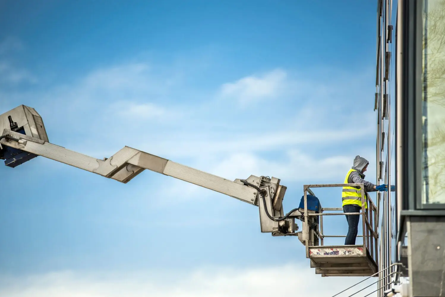 A person in a yellow safety vest and blue gloves stands in a boom lift platform, cleaning the glass facade of a building against a blue sky.