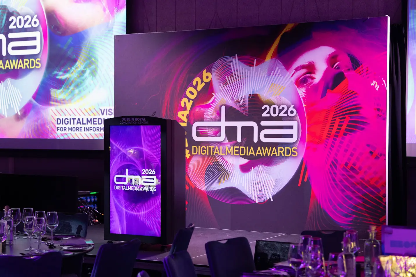 Event space with large screens displaying 'Digital Media Awards 2026' branding in vibrant pink, purple, and yellow. Tables with glasses are in the foreground.