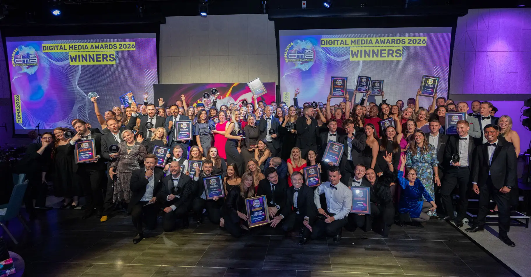 A large group of diverse people in formal wear celebrating and holding awards at the Digital Media Awards 2026, with 'WINNERS' displayed on screens behind them.