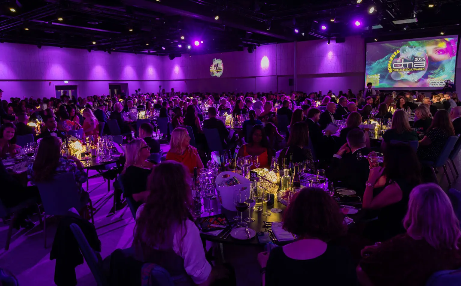 A large banquet hall filled with people seated at round tables, illuminated by purple lighting. A screen in the background displays 'DMA2026 Digital Media Awards'.