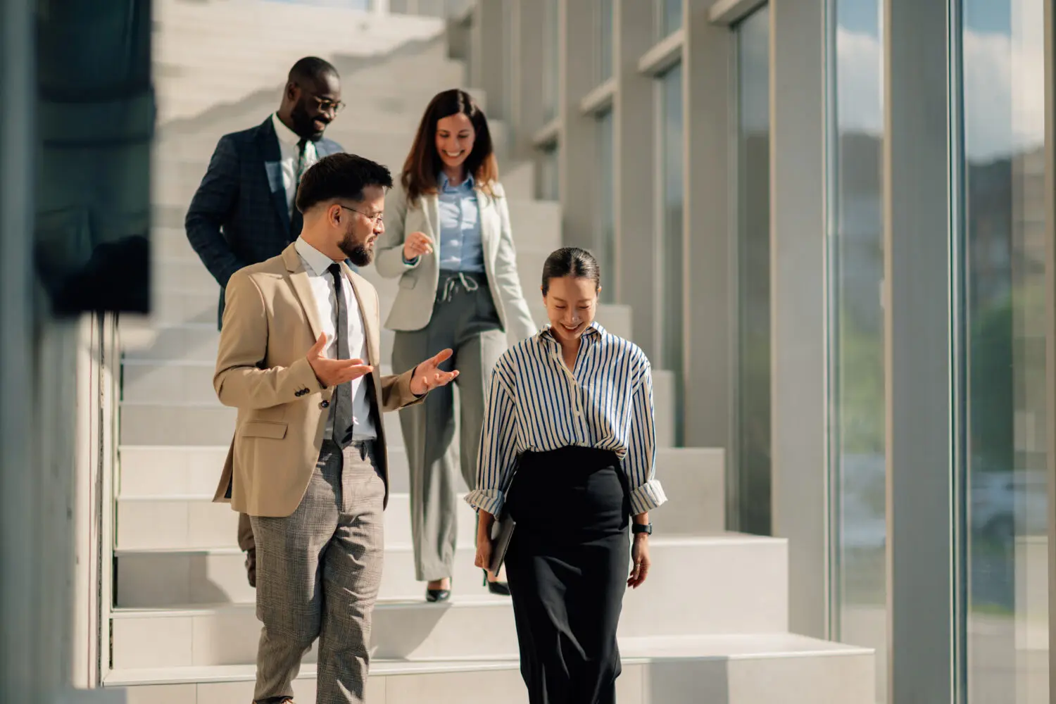 Four diverse business professionals, two men and two women, walk down a modern staircase, smiling and interacting. One man gestures while speaking to a woman holding a tablet.