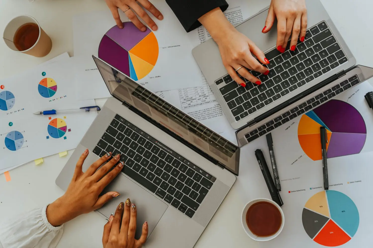 Overhead view of two people typing on laptops, surrounded by colorful pie charts, data reports, and coffee cups on a white desk.