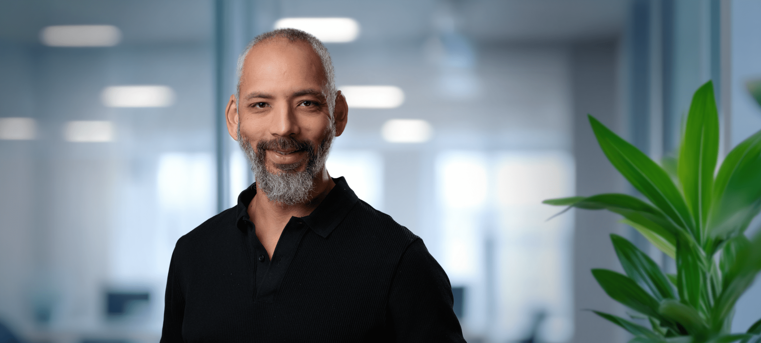 Headshot of a smiling man with a salt-and-pepper beard and black polo shirt, in a blurred office setting with a green plant.