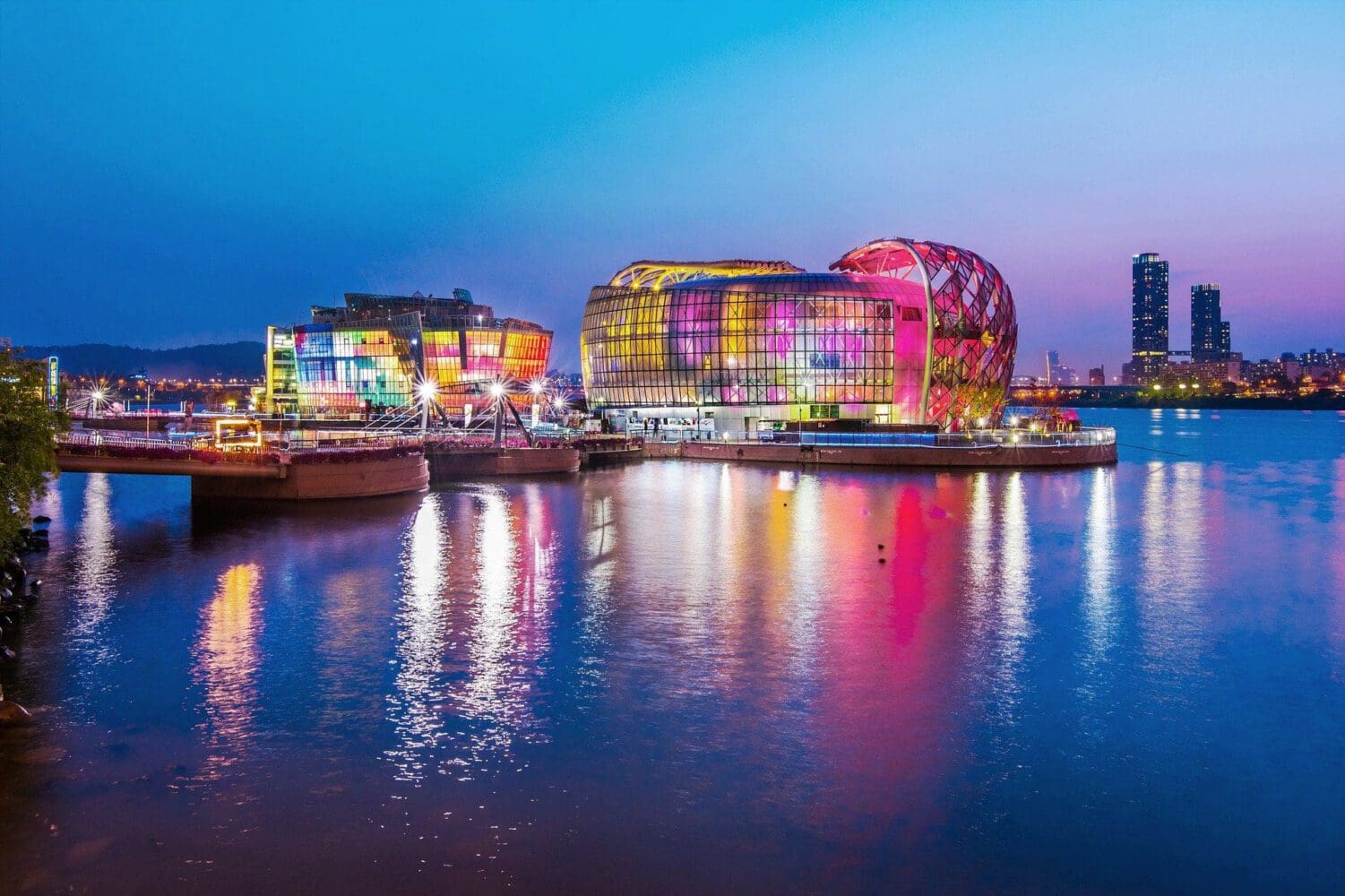 Three modern, glass-paneled floating structures on water, brightly lit in various colors like pink, yellow, and blue, reflecting on the dark water at night. A city skyline with skyscrapers is visible in the background.