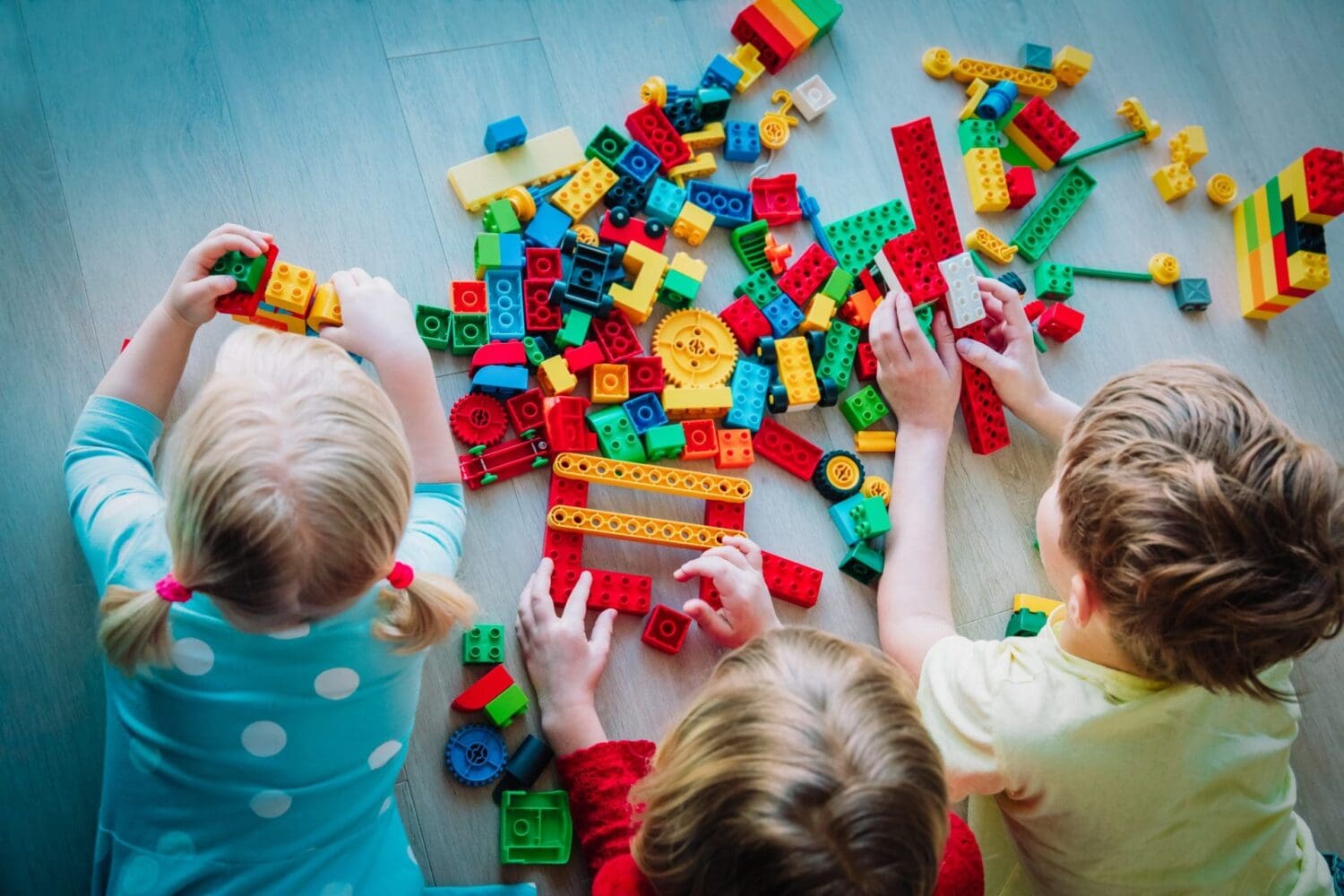 Three children playing with a large pile of colorful building blocks on a wooden floor, seen from an overhead perspective. Their hands are busy connecting various red, blue, green, and yellow blocks.