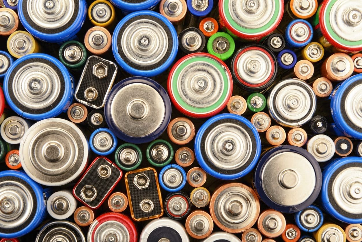 A close-up, top-down view of many different types and sizes of batteries, including cylindrical and 9-volt, in various colors like blue, red, green, and orange, tightly packed together.