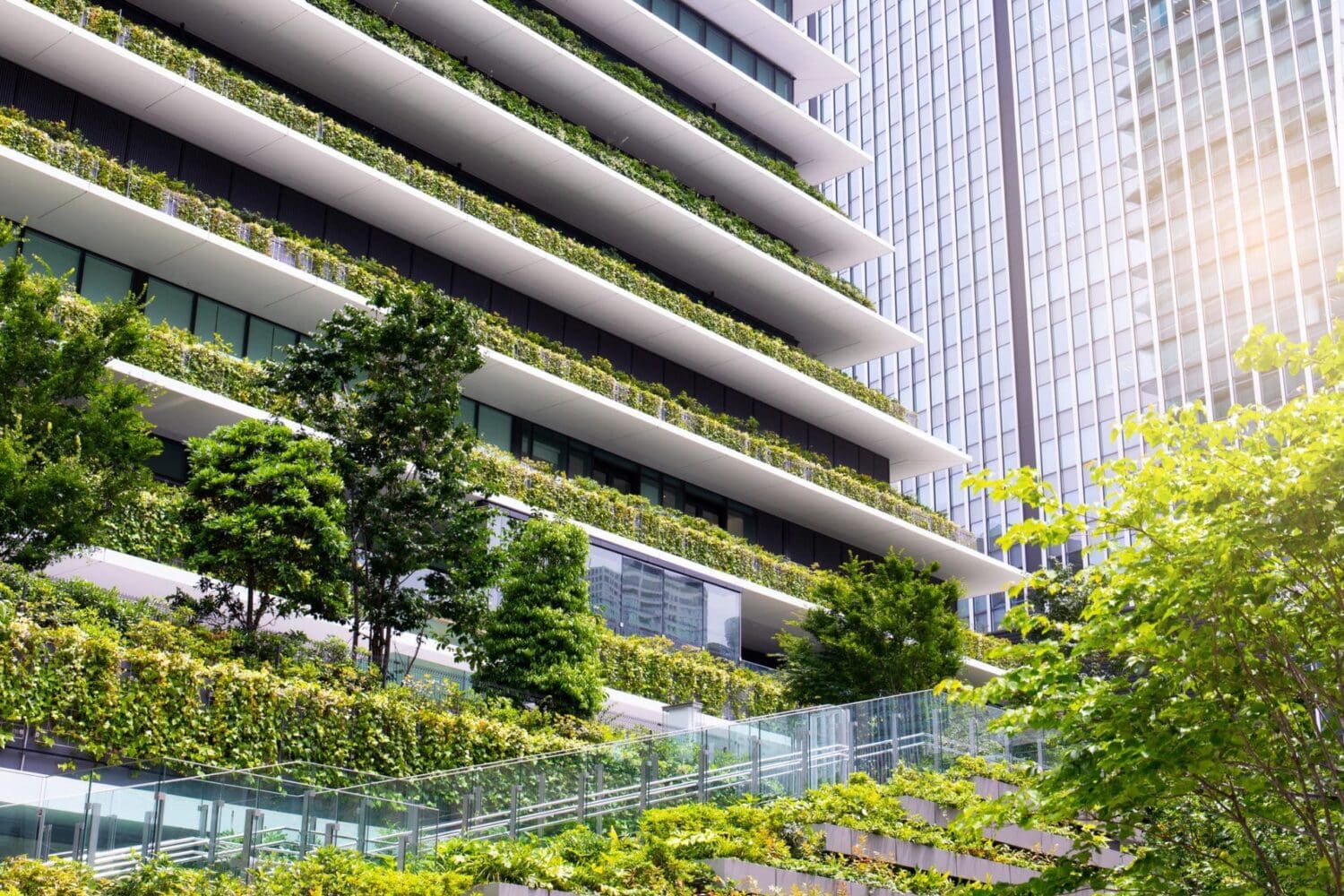 A modern building with terraced balconies covered in lush green plants and trees, next to a reflective glass skyscraper.