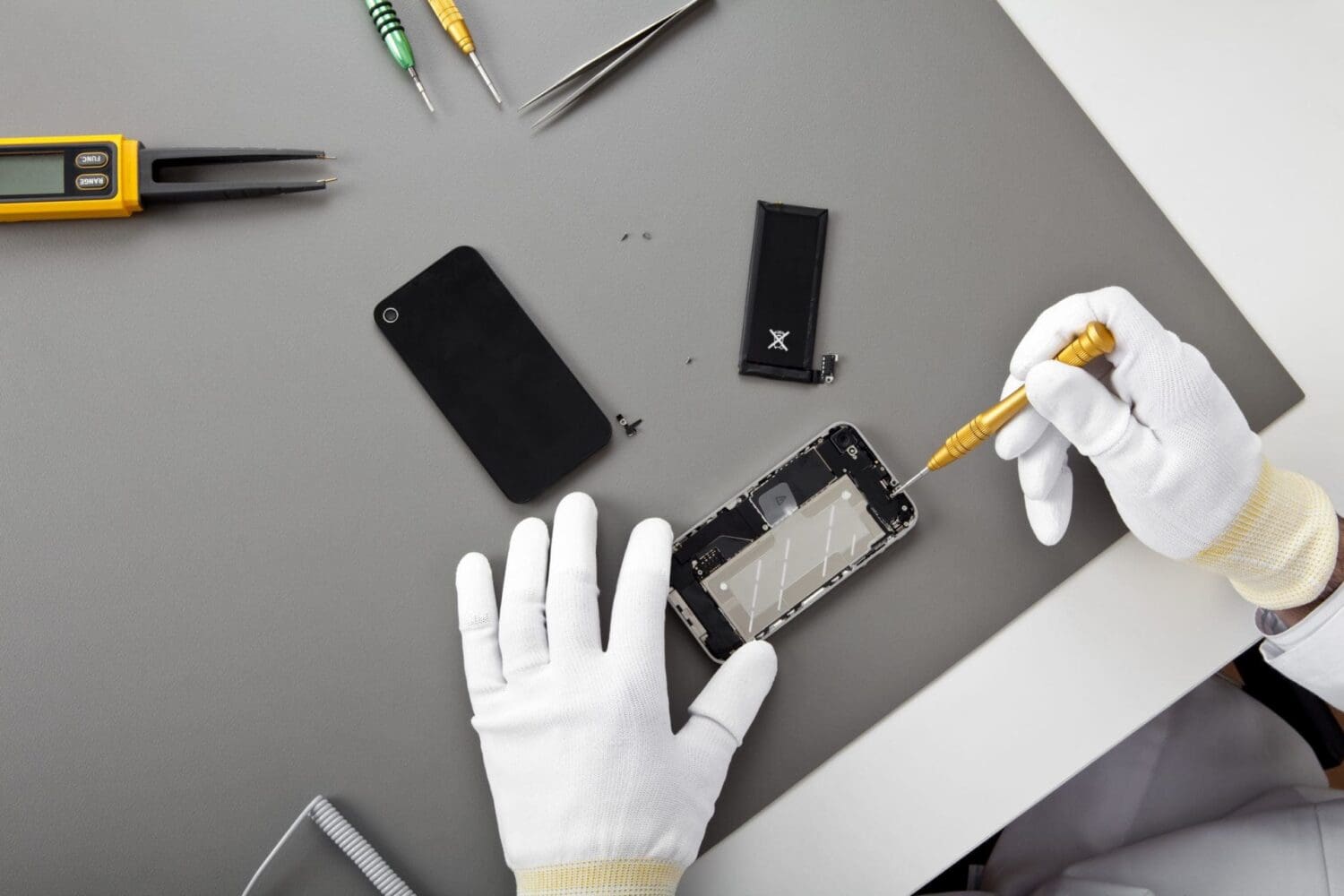 Overhead view of gloved hands repairing a smartphone on a gray surface, with tools and disassembled phone parts scattered around.