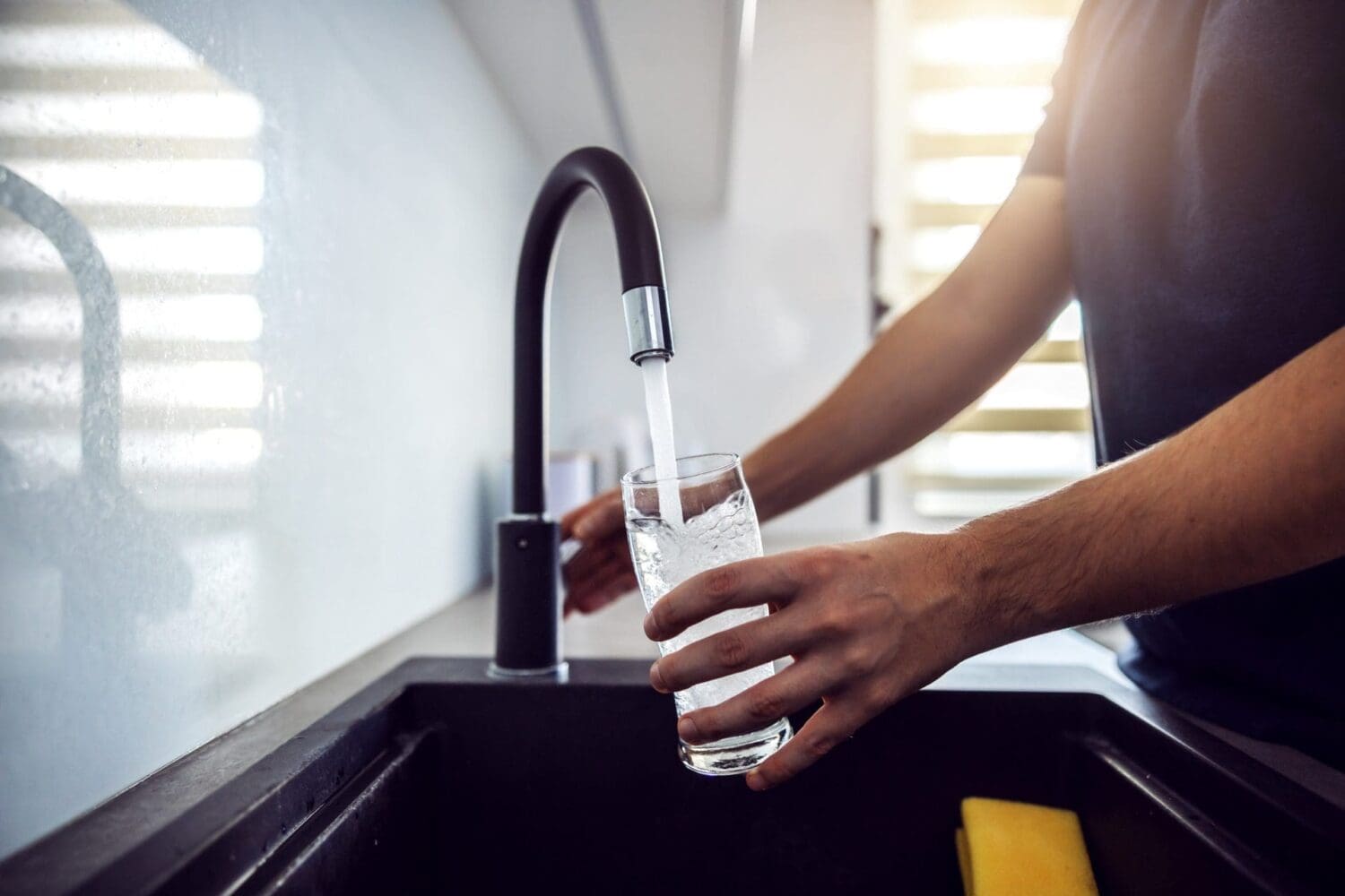 Close-up of hands filling a clear glass with water from a black kitchen faucet over a dark sink.