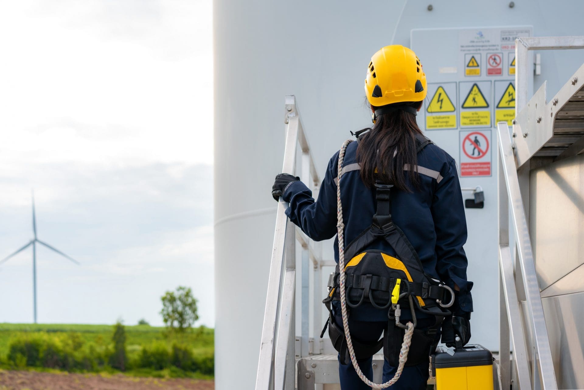A person in a yellow hard hat and safety harness stands on a metal ladder next to a wind turbine, holding a toolbox. Another wind turbine is in the distant background.