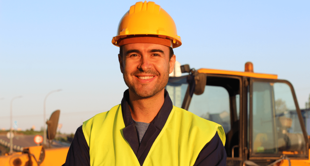 A smiling man in a yellow hard hat and high-visibility vest stands in front of heavy machinery.