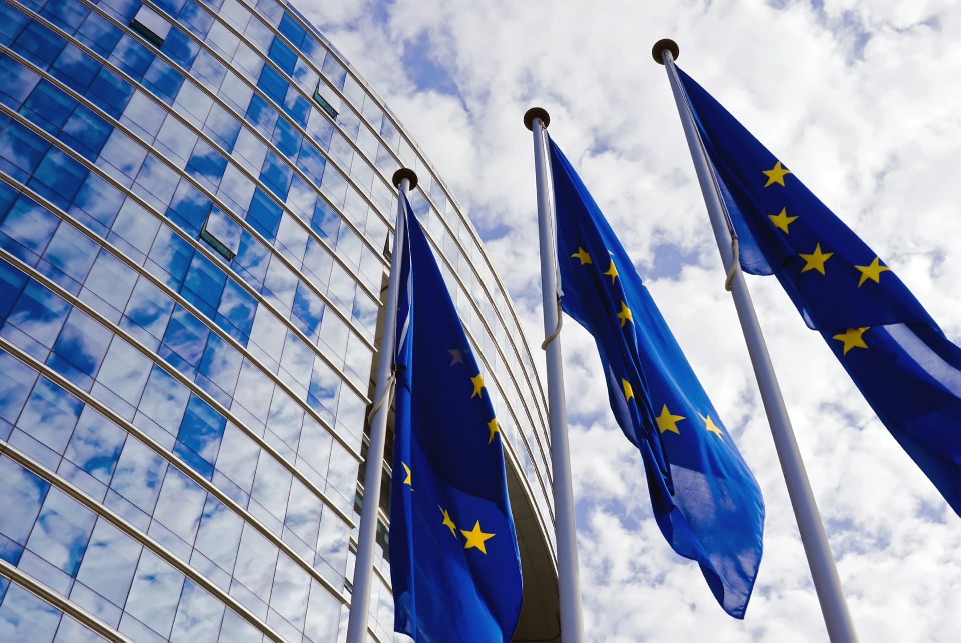 Three European Union flags waving in front of a glass building reflecting a cloudy sky.