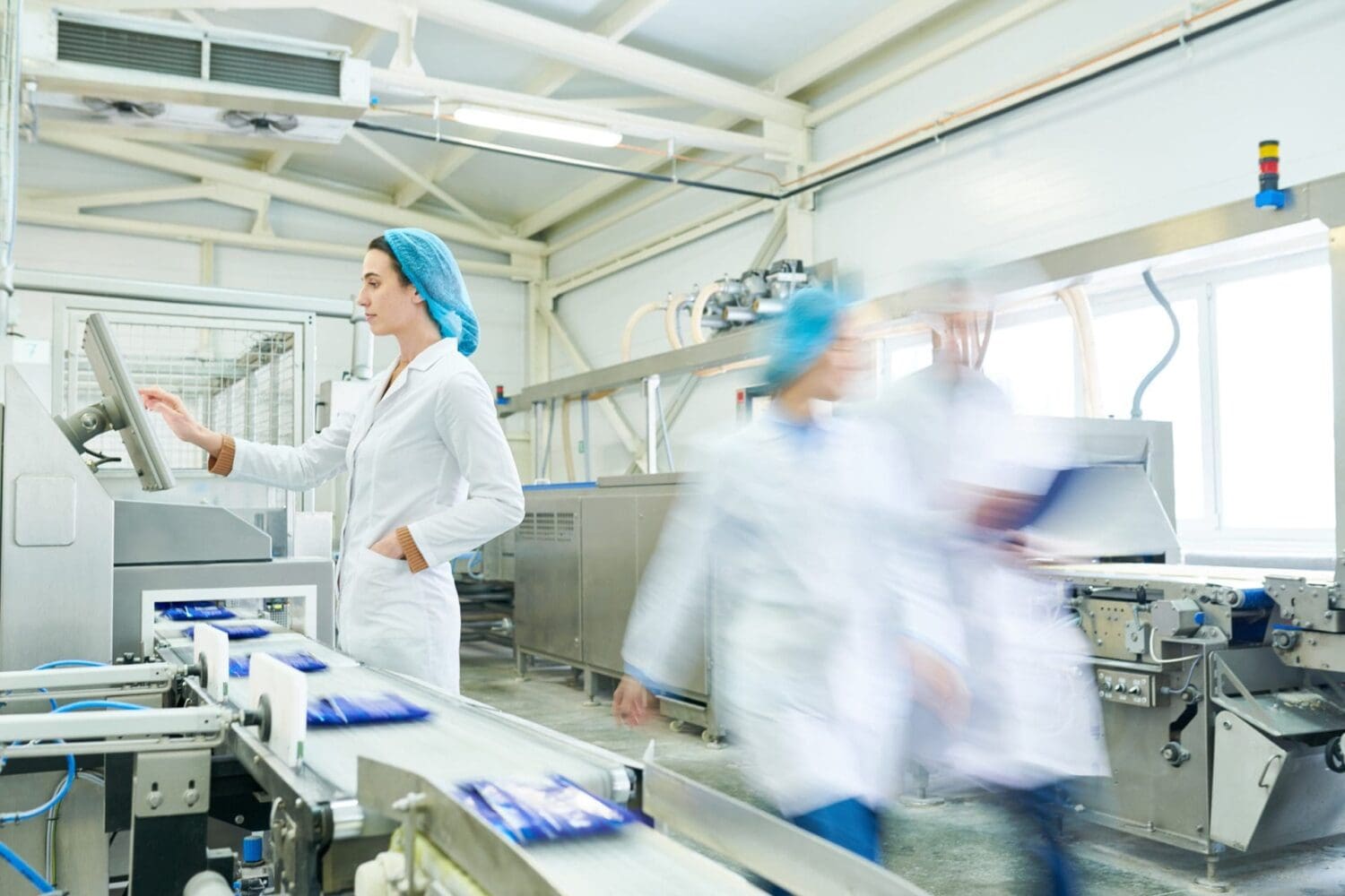 A woman in a lab coat and hairnet operates a touchscreen control panel next to a conveyor belt carrying packaged goods in a factory.