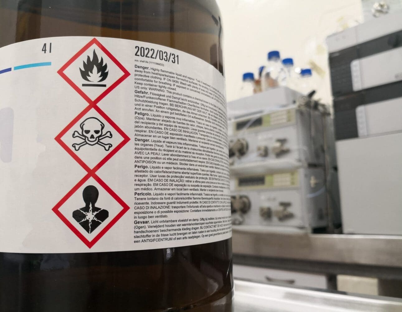 A brown chemical bottle with a white label showing GHS pictograms for flammability, toxicity, and health hazard, along with warning text and a shelf life date of 2022/03/31.