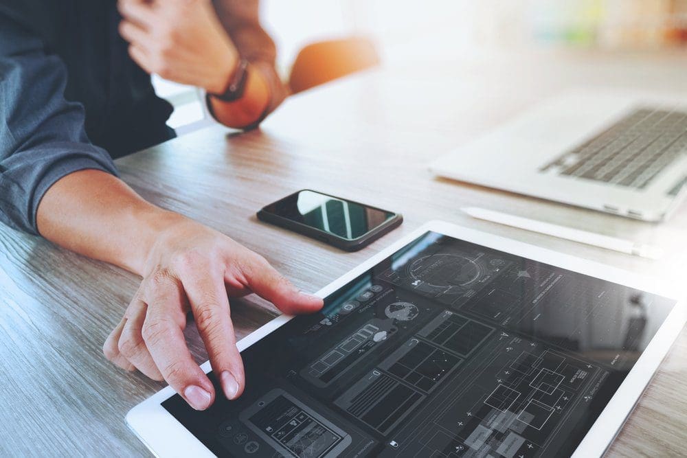 A person's hands interacting with a tablet displaying UI/UX design, a smartphone, and a laptop on a wooden desk.
