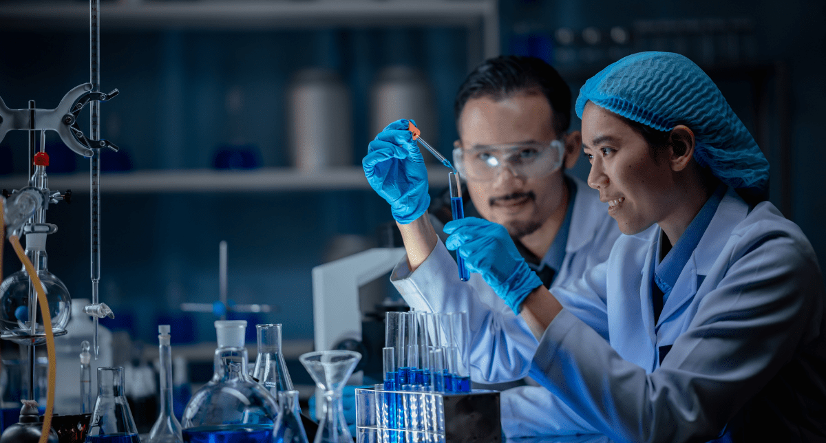 Two scientists in lab coats and blue gloves, one adding blue liquid to a test tube with a dropper while the other observes, in a laboratory.