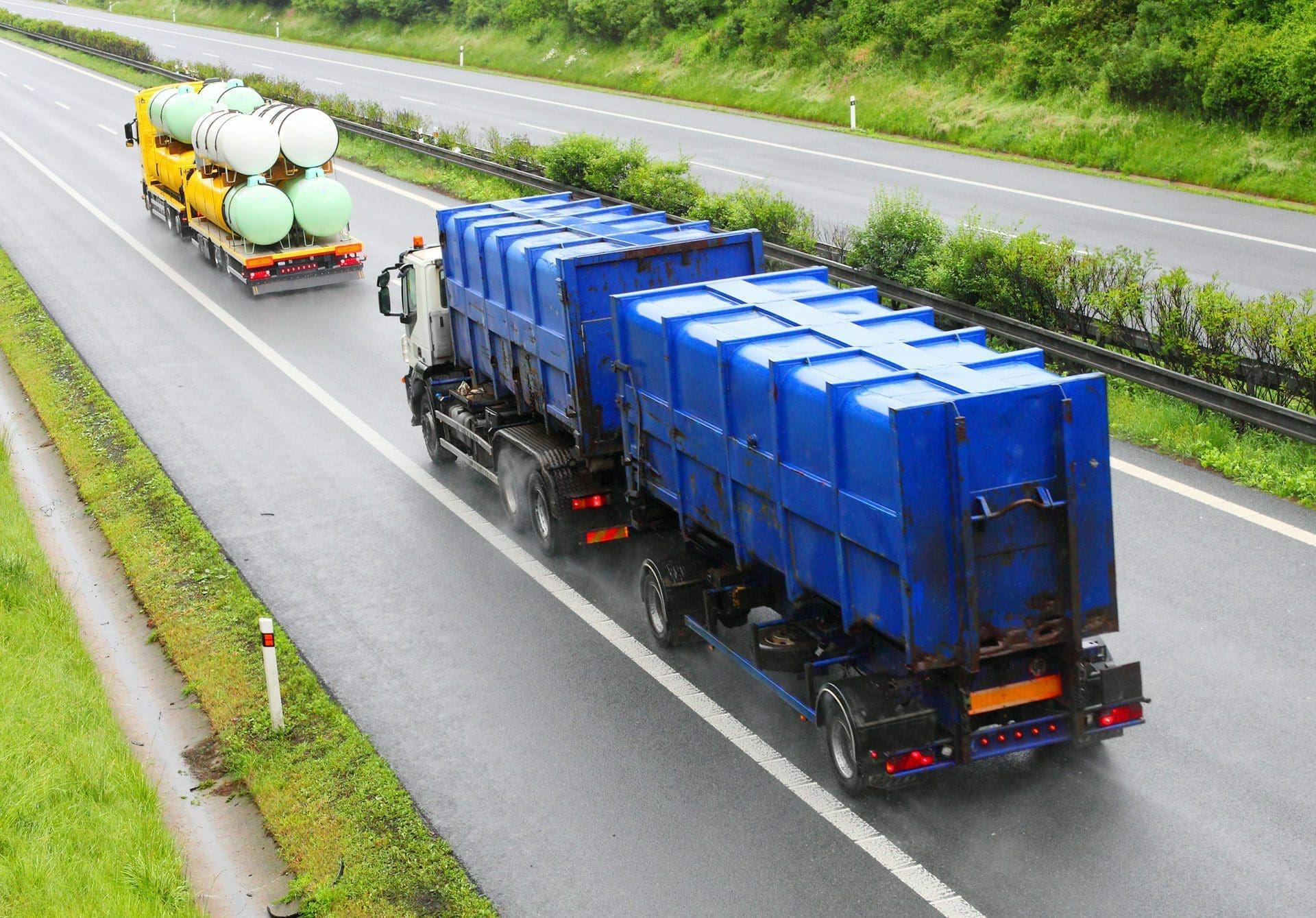 Two large trucks driving on a wet highway. One truck carries blue rectangular containers, and the other transports white and green cylindrical tanks.