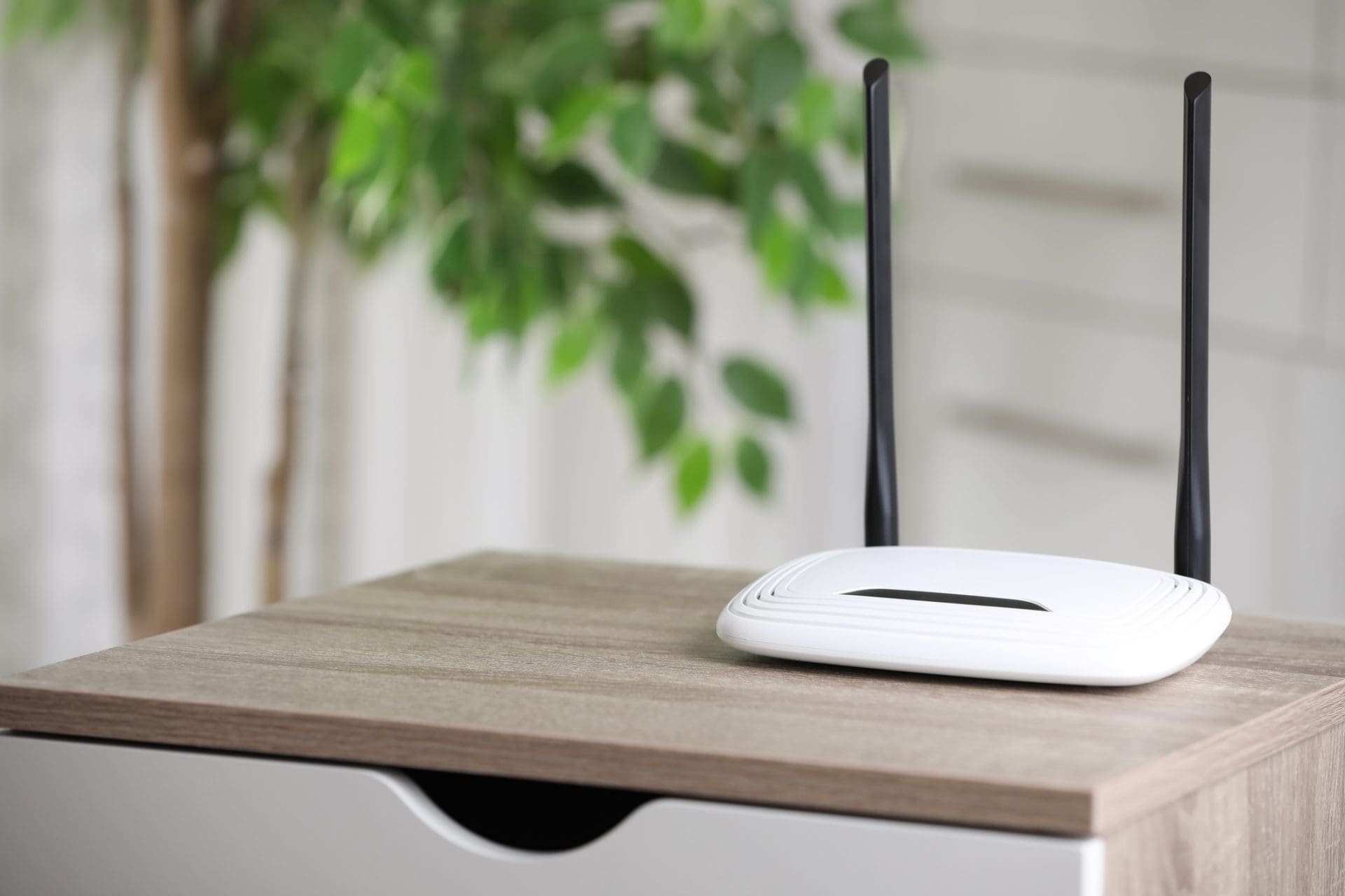 A white Wi-Fi router with two black antennas on a light brown wooden table, with a blurred green plant and light background.