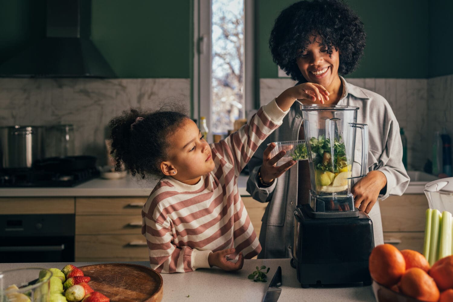 A smiling Black mother and her young daughter making a green smoothie in a kitchen. The daughter is adding greens to the blender, surrounded by fresh fruits like oranges, strawberries, and celery.