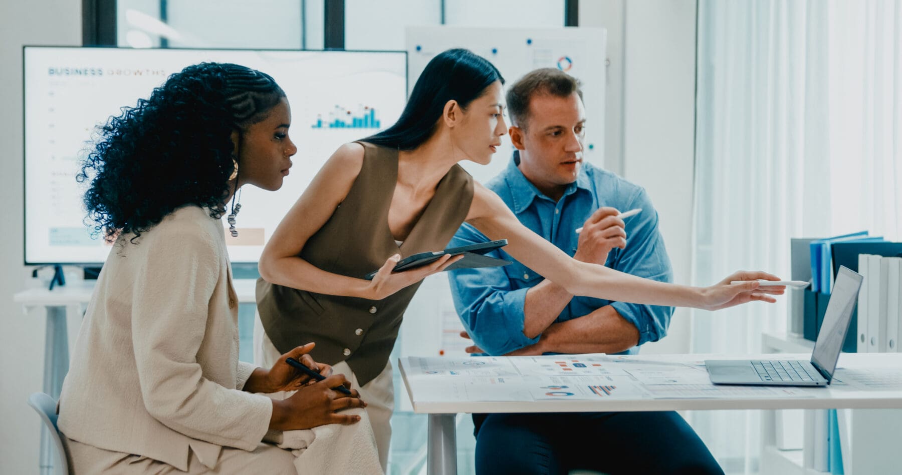 Three diverse professionals, two women and one man, are gathered around a laptop and documents in a bright office, discussing data displayed on a large screen in the background.