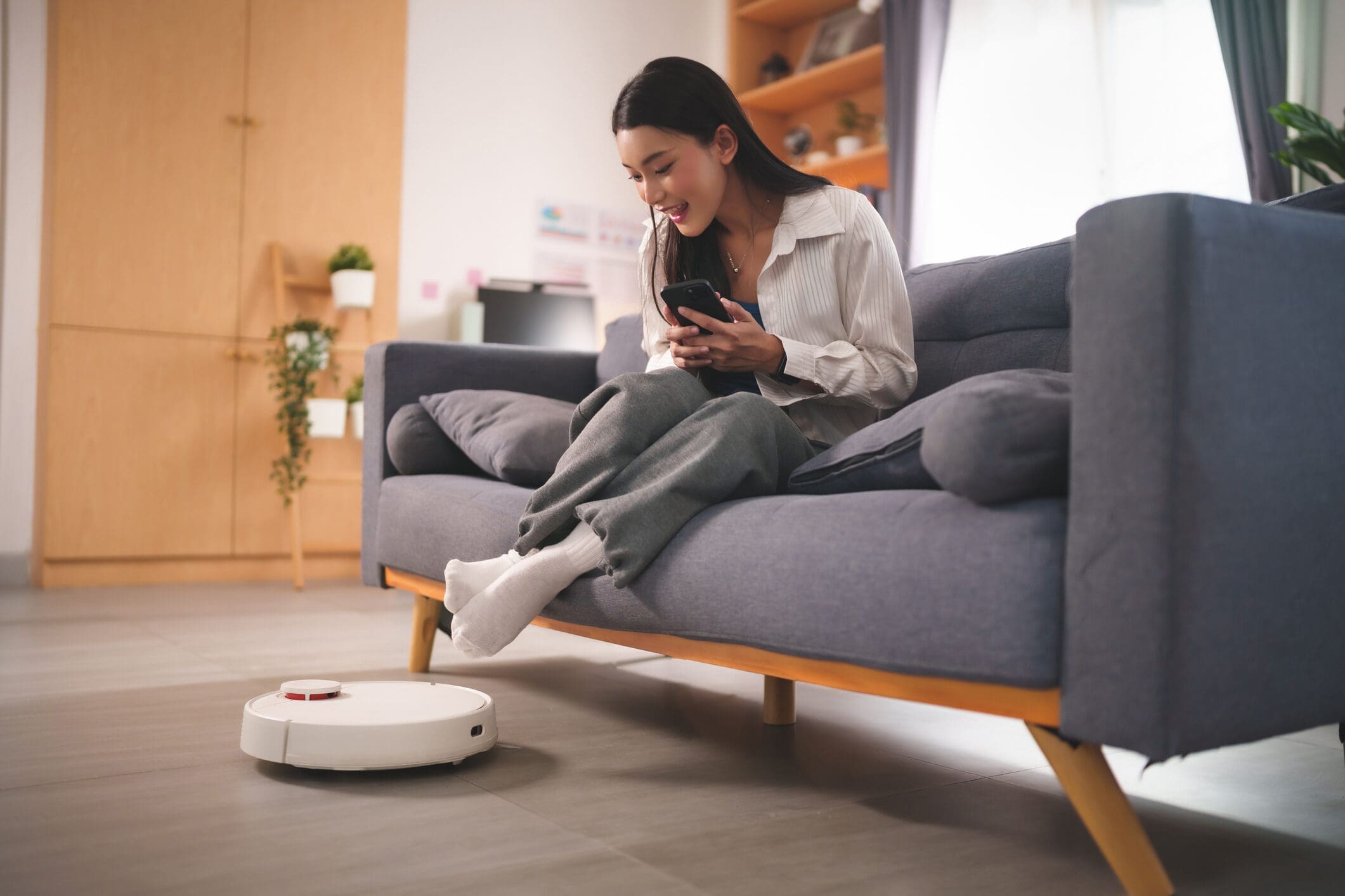 A young woman sits on a grey sofa, smiling at her phone, while a white robot vacuum cleans the floor in front of her.