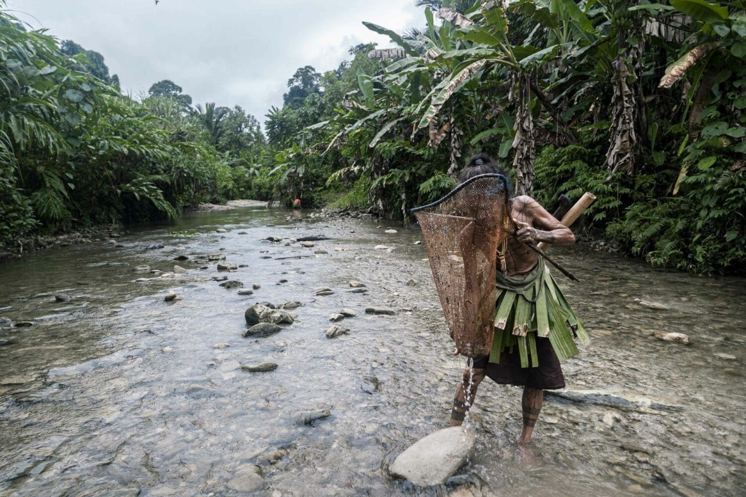 An indigenous person with tattoos and a leaf skirt wades through a rocky river, carrying a woven net basket and a wooden staff, surrounded by dense jungle.