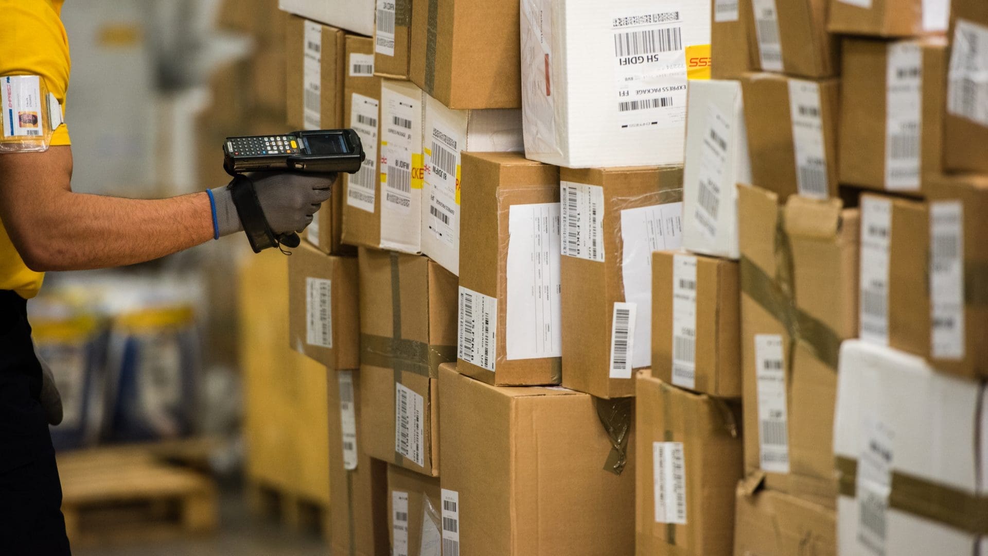 A person in a yellow shirt and grey glove uses a handheld barcode scanner to scan a tall stack of cardboard shipping boxes in a warehouse.