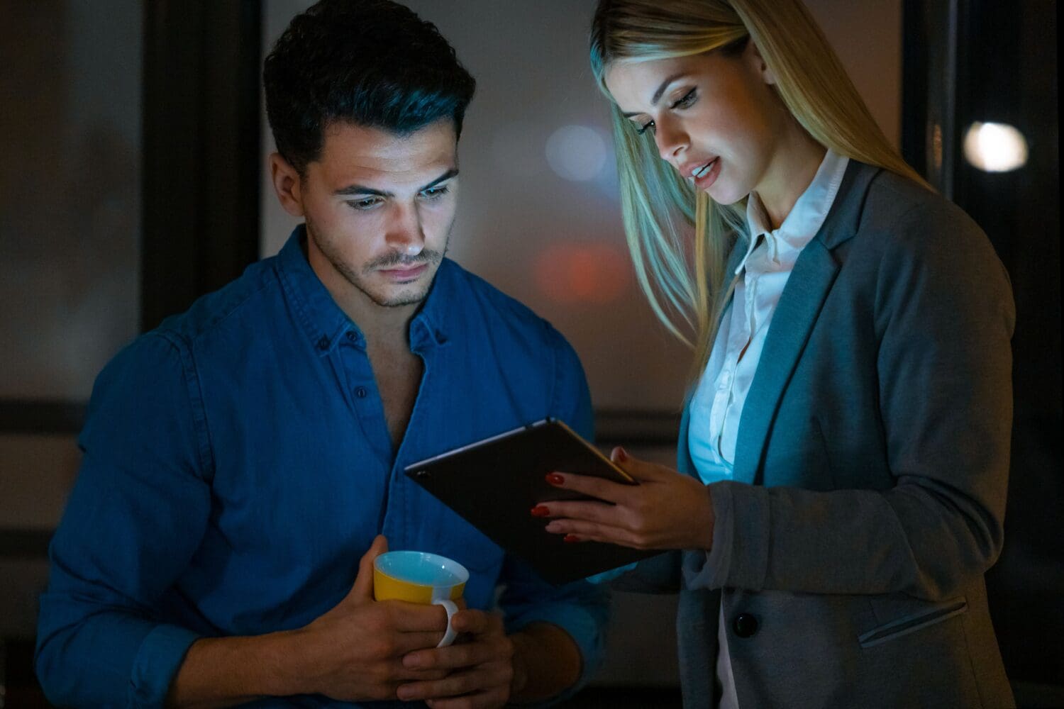 A man and a woman look at a tablet in a dimly lit room. The man holds a mug, and the woman holds the tablet.