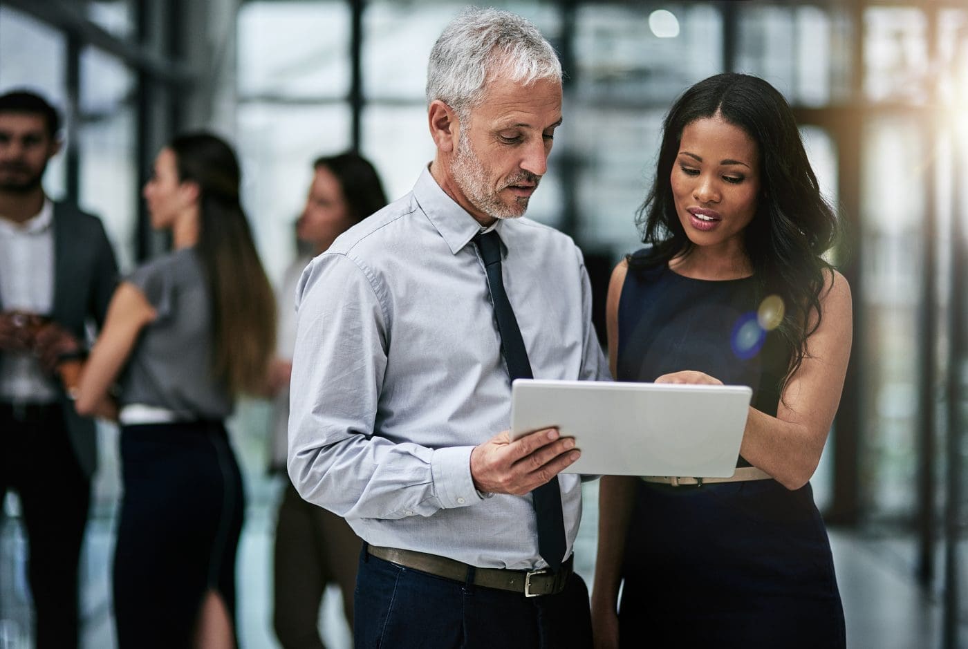 A man and a woman in an office setting look intently at a white tablet, appearing to discuss content on the screen. Other blurred colleagues are in the background.