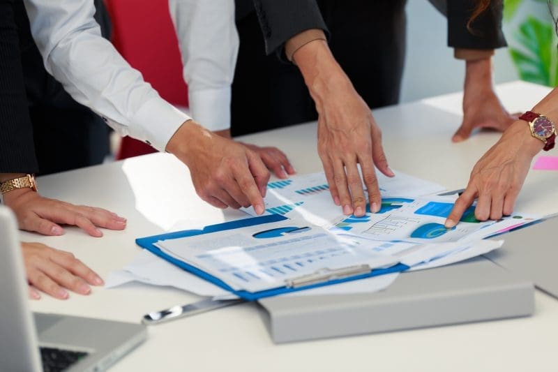 Close-up of multiple hands pointing at and discussing charts and graphs on documents spread across a white table, with a laptop nearby.