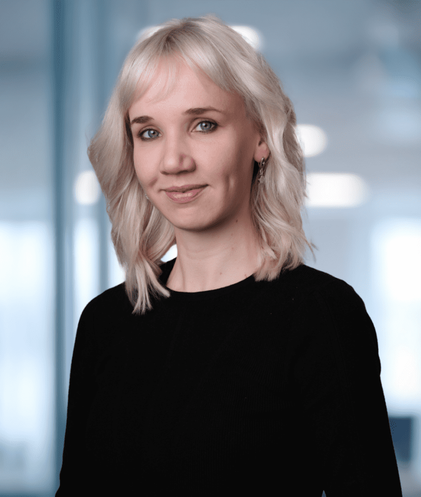 A smiling woman with shoulder-length blonde hair and a black top looks at the camera. The background is a blurred modern office with a green plant on the right.