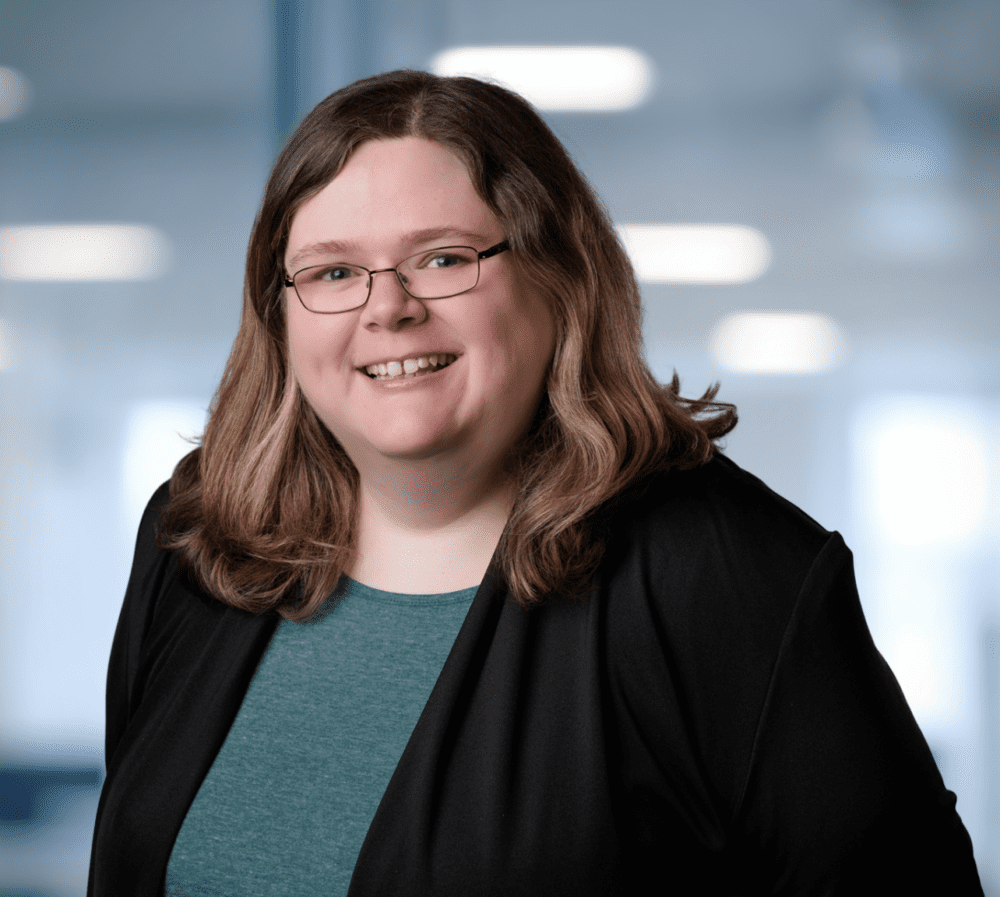 Headshot of a smiling woman with light brown hair and glasses, wearing a black cardigan over a teal top, in a blurred office setting with a plant.