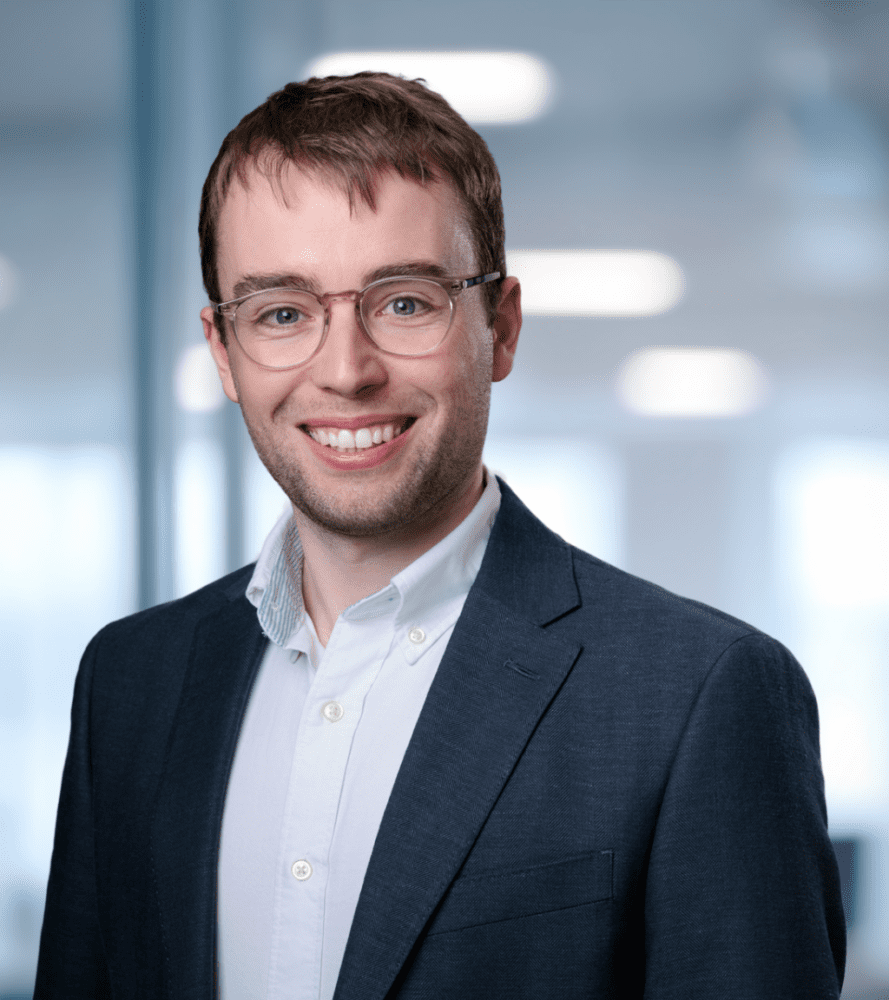 Headshot of a smiling young man with light brown hair and glasses, wearing a dark suit jacket and white shirt, in a blurred modern office setting with a green plant.