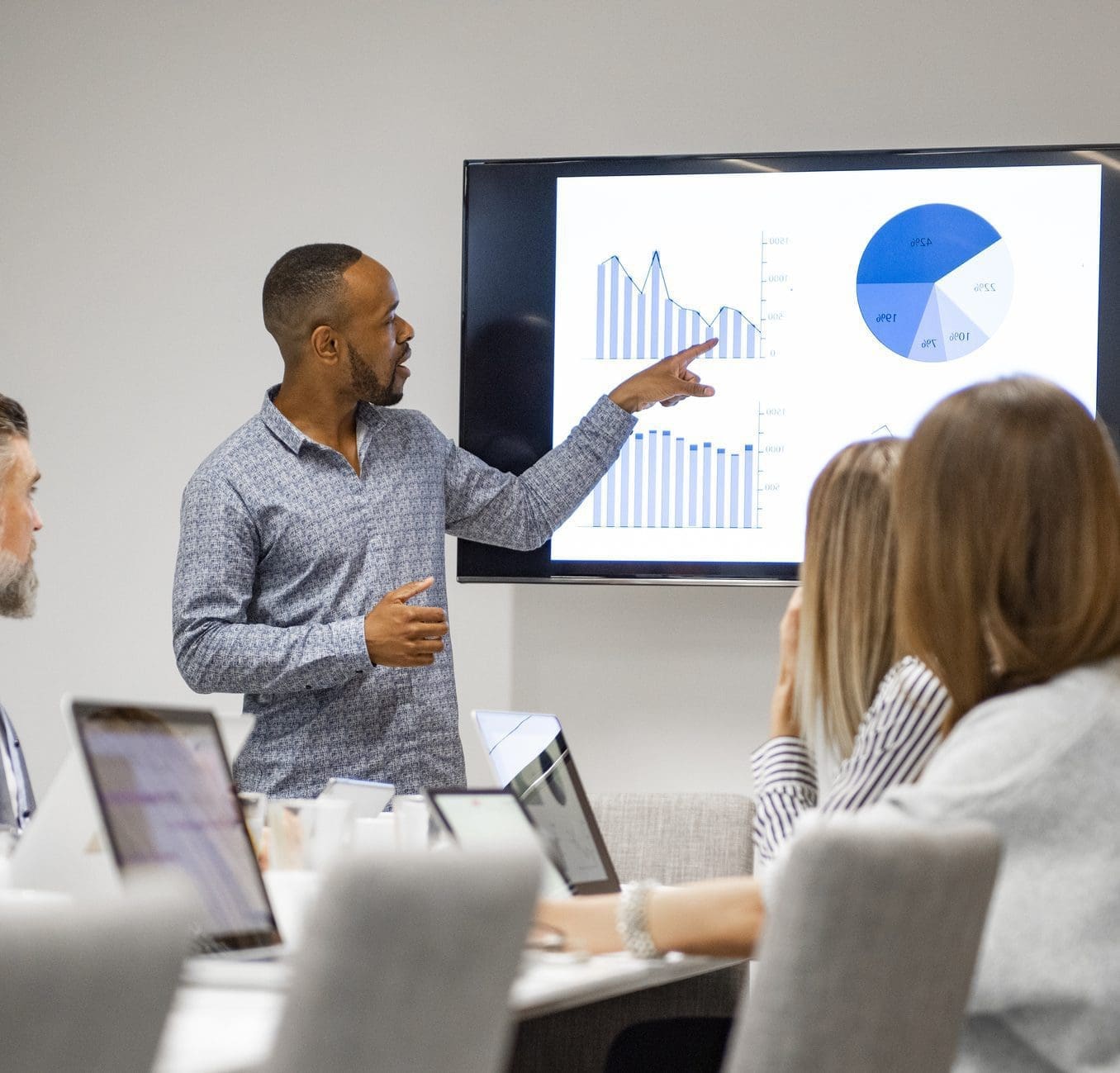 A Black man presents data visualizations on a large screen to a group of colleagues seated at a conference table.
