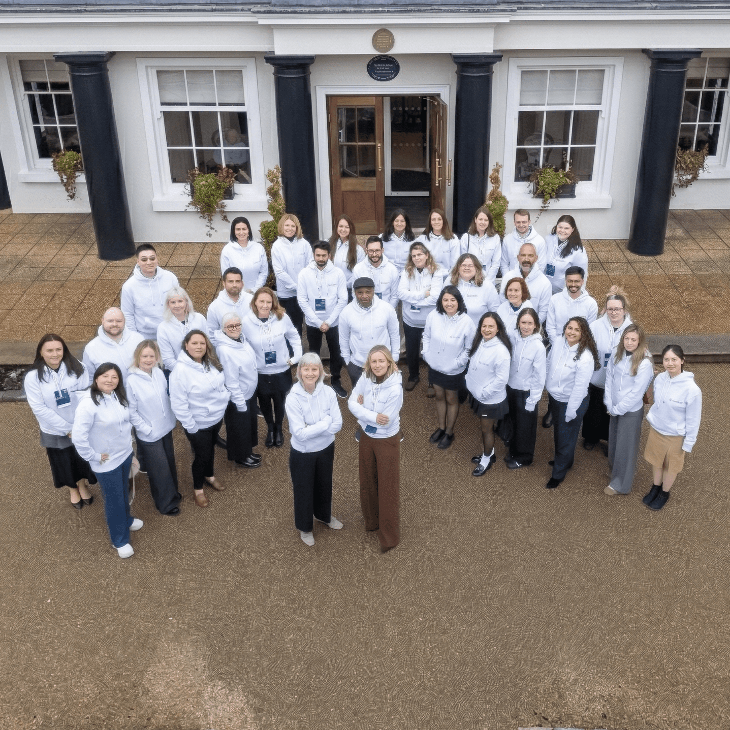 A high-angle photo of a large group of diverse people in matching white hoodies standing in a semi-circle in front of a grand white building with dark columns and a slate roof. Manicured hedges and paved ground are visible.