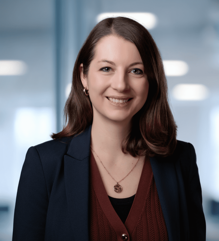 A smiling woman with dark brown hair wearing a navy blazer and a gold necklace, in a blurred office background with a green plant.