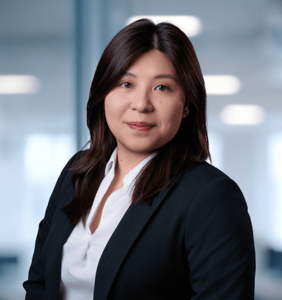 Professional headshot of an East Asian woman in a dark blazer and white shirt, smiling gently against a blurred office background.