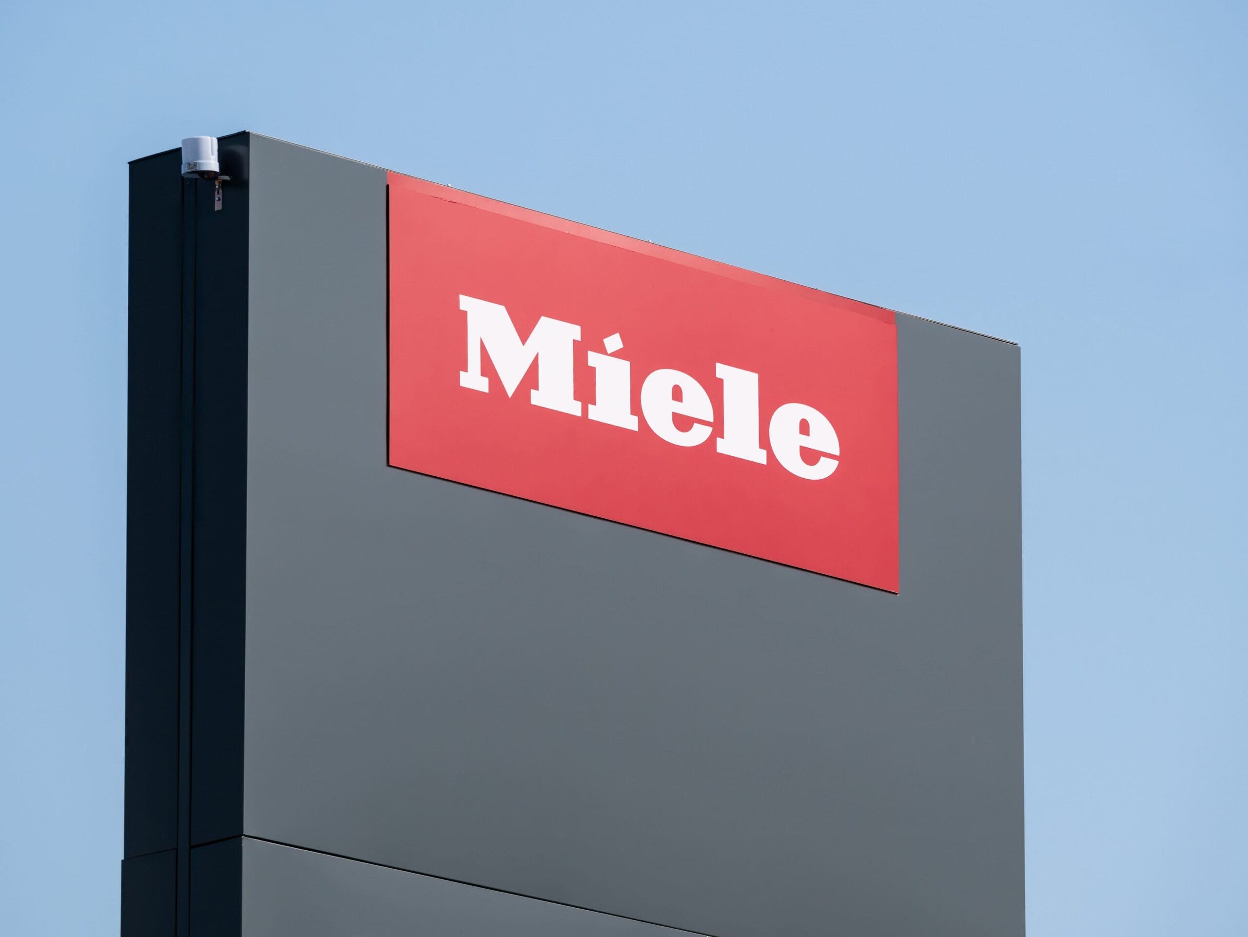 A Miele brand sign featuring the white 'Miele' logo on a red rectangle, set within a dark gray pylon, against a clear blue sky.