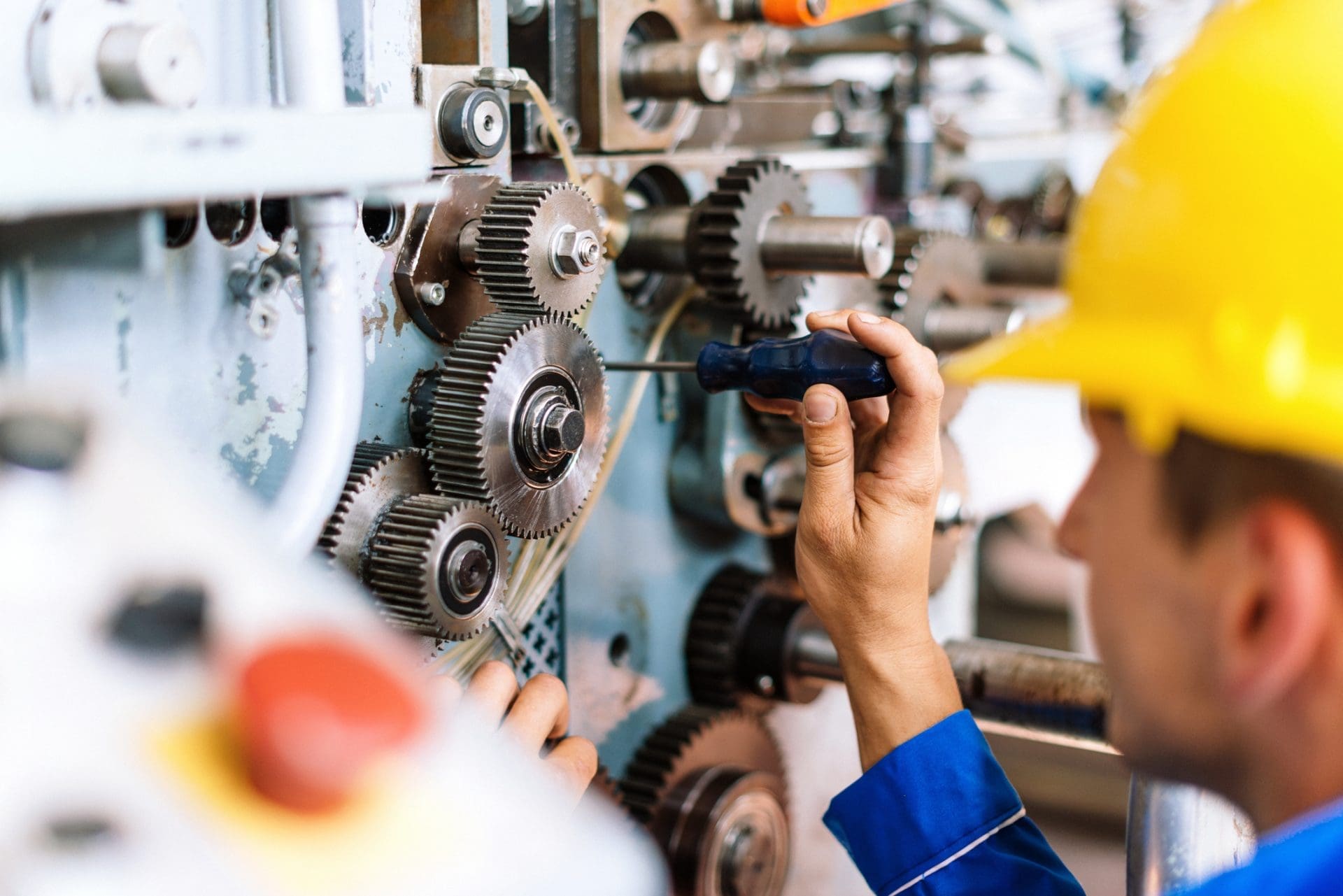 A person in a yellow hard hat and blue uniform uses a screwdriver to adjust metal gears on industrial machinery.
