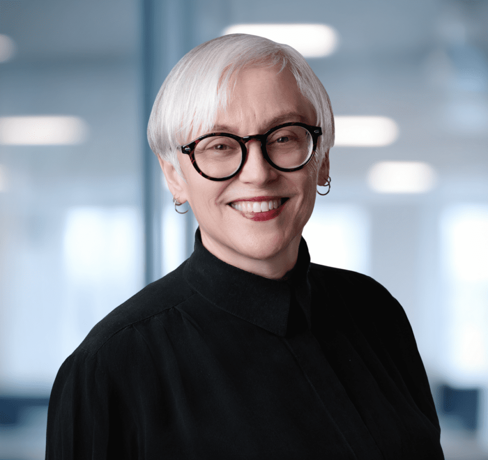 Headshot of a smiling woman with short white hair, round tortoiseshell glasses, and a black shirt, against a blurred modern office background with a green plant.